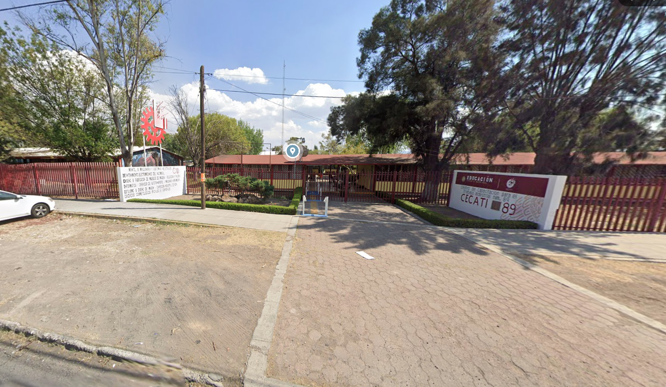 Fotografía en la que se muestra la entrada del plantel, conformada por una reja de color rojo que rodea las instalaciones. Del lado derecho hay un muro blanco y en letras rojas dice Cecati 89. Del lado izquierdo del muro se encuentra la reja que da acceso.